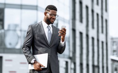 Takım elbiseli bir adam şık bir ofis binasının önünde duruyor, akıllı telefonuna gülücükle bakıyor. Kolunun altında bir dizüstü bilgisayar tutuyor. İşle ilgili faaliyetlerde bulunduğunu gösteriyor..