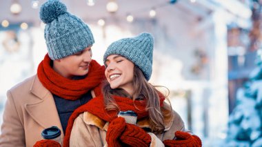 Sıcak şapkalar ve eşarplar takan neşeli bir çift kış harikalar diyarına yakın duruyor. Ellerinde sıcak içecekler, karın ve şenlikli dekorasyonların ortasında birbirlerine gülümsüyorlar..