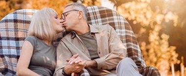 Yaşlı bir çift, büyük bir sandalyede birbirine yakın oturur ve sevgi dolu bir öpücüğü paylaşır. Gün batımının sıcak altın ışığı onları sarar, sakin bir açık hava sahnesinin ortasındaki neşeli anlarını vurgular..