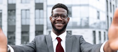 Takım elbiseli, kravatlı ve gözlüklü bir adam kentsel bir alanda durup hoş geldin jesti olarak kollarını uzatıyor. Arka planda parlak gün ışığında modern binalar yer alıyor.