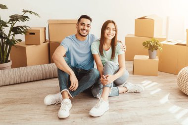 Mutlu bir çift yeni evlerinin zemininde oturuyor. Etrafı karton kutularla çevrili kameraya gülümsüyorlar..
