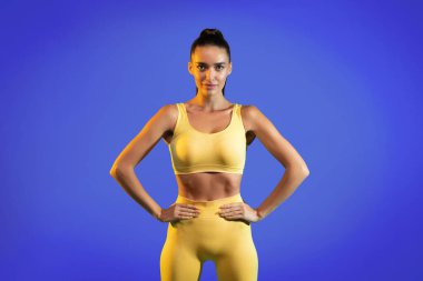 Formda bir model, elleri kalçasında, sarı bir spor kıyafeti sergiliyor. Arkaplan canlı bir mavi, enerjik bir atmosfer yaratıyor. Bu sahne fitness ve güveni vurguluyor..