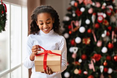 Heyecanlı Afrikalı Amerikalı kız, Noel 'i Noel neşesiyle dolu dekore edilmiş bir oturma odasında kutlarken kırmızı kurdeleli hediye kutusunu açıyor. O anda saf bir sevinç gösteriyor..