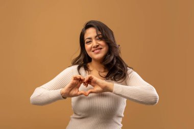 Dost canlısı Arap kadın kalp şeklinde eller, göğse yakın aşk jestleri yapıyor, kameraya gülümsüyor, stüdyoda kahverengi arka planda poz veriyor.