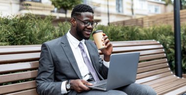 Takım elbiseli genç bir adam parkta bir bankta oturuyor, kahvesini yudumlarken dizüstü bilgisayarını kullanıyor. Sahne, yeşillik, verimlilik ve gevşeme içeren canlı ve sıcak bir günü yakalıyor..