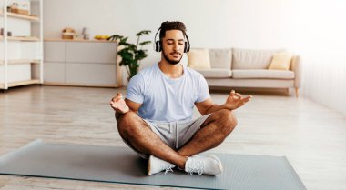 Genç bir adam, parlak, dingin bir odada yoga minderinde bağdaş kurmuş, kulaklık takmış ve meditasyona odaklanmış oturuyor. Yumuşak doğal ışık boşluğu doldurur, barışçıl bir atmosfer yaratır..