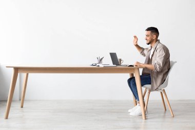 Genç bir Arap, parlak bir ofisin ahşap masasında oturmuş bilgisayarında video görüşmesi yapıyor. Gülümsüyor ve web konferansı sırasında kameraya el sallıyor. Bağlılık ve samimiyet gösteriyor..