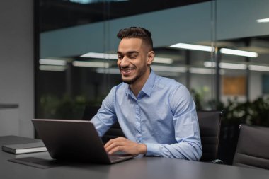 Neşeli genç bir ortadoğulu yönetici, e-posta gönderirken ve pazarlama araştırması yaparken, modern bir ofiste, bilgisayarına odaklanmış bir şekilde bir çalışma masasında oturuyor. Ofisin rahat bir atmosferi var..