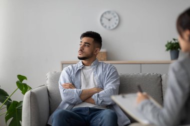 Danışmanlık bürosunda genç bir Arap, düşünceli ve mesafeli bir şekilde bir koltuğa oturur. Bir psikolog, zihinsel sağlığa odaklı profesyonel bir çevrede destek sağlamak için notlar alıyor..
