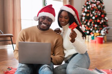 Gülümseyen siyah çift oturma odalarında halının üzerinde oturmuş internetten alışveriş yapıyorlar. Noel süsleri ve şenlikli ağaçlarla çevrili bir kredi kartı gösteriyorlar..