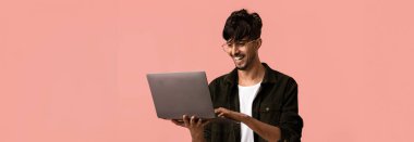 Genç bir adam pembe arka planda dizüstü bilgisayar kullanırken gülümsüyor. Günlük kıyafetler ve gözlük takıyor, göreviyle nişanlandığını gösteriyor..