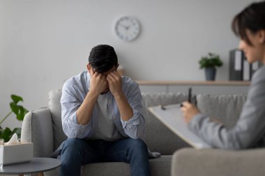 Orta Doğulu bir adam bir kanepeye oturur, kafasını stres içinde tutarken bir psikolog profesyonel bir ortamda dikkatle dinler. Oturumda kişisel zorluklar ve duygusal iyileşmeler ele alınıyor..