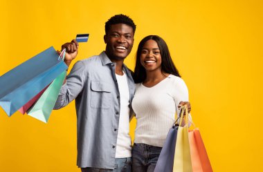 Neşeli bir çift, ellerinde çeşitli renklerde alışveriş torbaları ve kredi kartı olan, parlak sarı bir arka plana karşı birlikte durarak başarılı bir alışveriş gününü kutluyor..