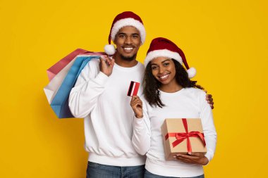 Mutlu bir çift, ellerinde alışveriş poşetleri ve hediye kutusu olan şenlikli bir kıyafet giyer. Parlak sarı bir arka plana gülümsüyorlar, bayram alışverişinin ve kutlamanın ruhunu yakalıyorlar..