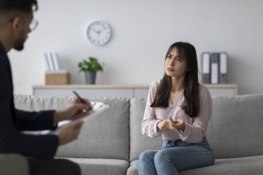 Üzgün genç bir Arap kadın klinikteki bir kanepede oturuyor ve bir danışma sırasında duygularını bir psikologla paylaşıyor. Bu toplantıda zihinsel sağlık desteği ve anlayış ele alınıyor..