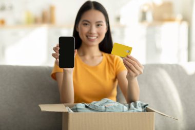 Asyalı kadın elinde karton bir kutuyla kanepeye oturmuş elinde bir telefon ve kredi kartı tutarken gülümsüyor. Konforlu bir ev ortamında modern alışveriş ve internet bankacılığını kucaklıyor..