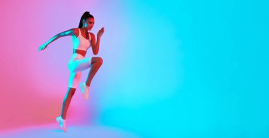 Atletik giyinen bir kadın canlı pembe ve mavi ışıklandırmalı bir stüdyoda sıçrama yapar. Enerjisini ve gücünü gösteriyor, aktif ve sağlıklı bir yaşam tarzının altını çiziyor..