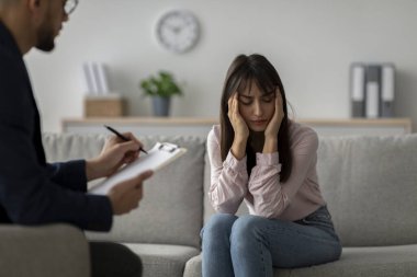 Arap kökenli bir kadın, psikolog muayenehanesinde koltukta görünür bir şekilde sıkıntılı bir şekilde oturuyor. Terapi seansı sırasında şakaklarına dokunuyor, stres belirtileri gösteriyor. Terapisti dikkatle dinliyor..