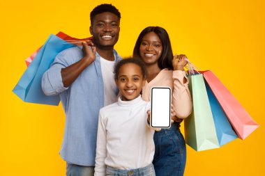 Neşeli siyah bir aile, parlak sarı bir stüdyoda ellerinde alışveriş torbalarıyla bir arada duruyor. Bir kız gururla boş bir akıllı telefon ekranı sergiliyor, online alışveriş promosyonları için mükemmel..