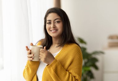 Neşeli genç bir ortadoğulu kadın elinde sıcak bir bardak içecekle pencerenin önünde duruyor. Evde dinlendirici bir hafta sonu sabahında kameraya gülümsüyor ve kahve molasının tadını çıkarıyor..