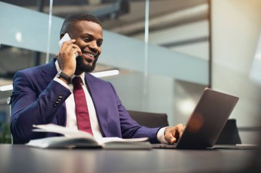 Pahalı mavi takım elbiseli başarılı genç iş adamı ofiste dizüstü bilgisayarda çalışıyor, kişisel asistanı ya da iş ortağıyla telefon görüşmesi yapıyor ve gülümsüyor.