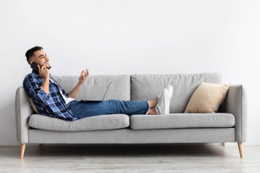 Gülümseyen bir Arap, rahat oturma odasında rahat bir kanepede uzanıyor ve akıllı telefonuyla konuşuyor. Konuşurken el kol hareketi yaptığı için meşgul ve mutlu görünüyor..