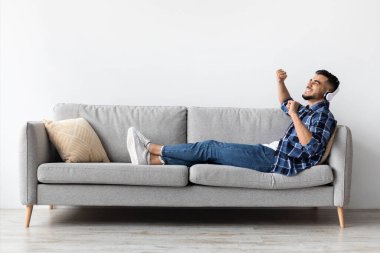 Neşeli Orta Doğulu bir adam rahat bir kanepeye oturur, şarkı söylerken ve dans ederken kulaklık takar. Keyifli bir oturma odasında dinlenip müzik dinlemeyi seviyor..