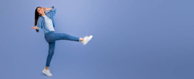 Bir kadın dans ederken gülümser, bir bacağını havaya kaldırır. Kotlardan ve beyaz spor ayakkabılardan oluşan sıradan bir kıyafet giyiyor. Saf mavi bir arka planda canlı bir sahne yaratıyor..