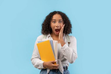 Kıvırcık saçlı kız öğrenci şok olmuş ve korkmuş görünüyor, elinde defterler ve sırt çantası var. Mavi bir arkaplanda yapılacak üniversite sınavından duyduğu endişeyi dile getiriyor..