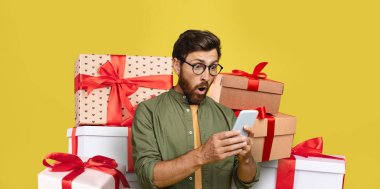 Renkli hediye kutularının önünde kurdeleli bir adam duruyor. Telefonunu kullanırken heyecanlı bir bakış atıyor. Parlak sarı arka plan, sahnenin kutlama havasını arttırır..