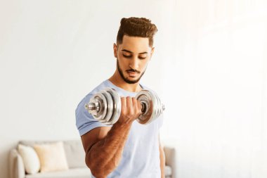 A fit man is lifting a dumbbell in a well-lit room, focusing on his workout. He is dedicated to building strength and improving his fitness during this training session.