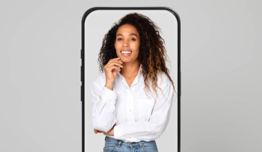 Kıvırcık saçlı genç bir kadın, kendinden emin bir şekilde ayakta dururken neşeli bir ifade sergiler..