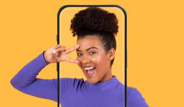 Neşeli genç bir kadın, canlı sarı bir arkaplan önünde neşeli bir poz vererek eliyle barış işareti yapıyor. Bir telefon çerçevesi tutuyor ve sahneye yaratıcılığı ekliyor..