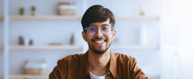 Gözlüklü neşeli bir genç adam modern bir odada oturuyor ve kameraya gülümsüyor. Mekan şık mobilya ve bitkilerle aydınlık, hoş bir atmosfer yaratıyor..