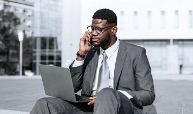 Takım elbiseli bir adam dışarıda yerde oturuyor, dizüstü bilgisayarla çalışırken telefonla konuşuyor. Arka plandaki modern bina yoğun bir şehir ortamını yansıtır..