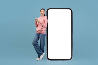 Genç bir Çinli kadın, boş bir akıllı telefon ekranına yaslanırken gülümsüyor. Daha küçük bir akıllı telefonu var, mobil uygulamalarla meşgul..