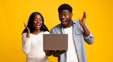 İki arkadaş heyecanla gülümsüyor ve dizüstü bilgisayar ekranına bakarken ellerini kaldırıyorlar. Parlak sarı arka plan neşeli anlarına neşeli bir atmosfer ekler..