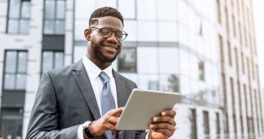 Genç bir işadamı şık bir ofis binasının önünde mutlu mesut tabletle meşgul olarak duruyor. Takım elbise ve gözlük takıyor, hareketli bir şehir ortamında işinden zevk alıyor..