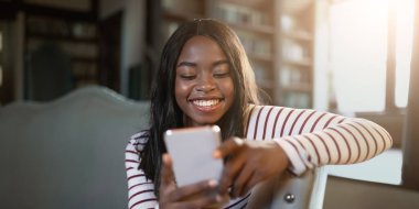 Bir kadın parlak bir odada rahatça oturur, akıllı telefonunu kullanırken gülümser. Ilık ışık etrafında hoş bir atmosfer yaratıyor ve neşeli ifadesini arttırıyor..