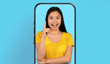 Genç bir kadın işaret parmağını telefon çerçevesinin içinde tutarken gülümsüyor. Sarı bir gömlek giyiyor ve parlak mavi bir arka plana karşı duruyor, ilgi çekici ve eğlenceli bir sahne yaratıyor..