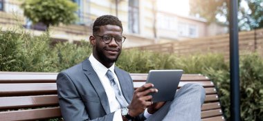 Gri takım elbiseli genç bir adam parktaki bankta rahatça oturuyor. Yeşil bitkiler ve sıcak bir atmosferle çevrili güneşli öğleden sonranın tadını çıkarırken bir tablet kullanır..