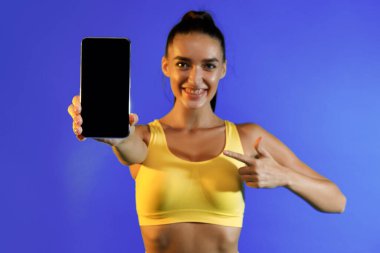 Sarı bluzlu bir kadın, parlak mavi bir arka plana karşı akıllı telefonu işaret ederken gülümsüyor. Bu, bir fitness bağlamında coşku ve bağlantı anını yakalar.