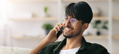 Gözlüklü genç bir adam evinde rahat bir şekilde oturur ve sıcak bir gülümsemeyle telefonda sohbet eder. Parlak odada rahat bir havayı yansıtan bitkiler ve rahat bir atmosfer var..