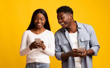 A young couple stands together, both looking at their smartphones with smiles. They seem to enjoy a lighthearted moment, sharing laughs against a cheerful yellow backdrop.