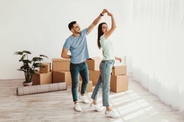 Bir çift yeni evlerinde paketlenmemiş kutularla dans ederken eğlenceli bir anın tadını çıkarıyor. Güneş ışığı pencerelerden içeri akarak sıcak ve davetkar bir atmosfer yaratır..