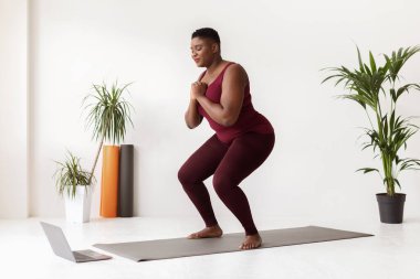 Genç ve iri yarı bir zenci kadın evde spor paspası üzerinde egzersiz yapıyor. Dizüstü bilgisayarında egzersiz videosu izlerken çömeliyor, sağlığına ve sağlığına olan bağlılığını gösteriyor..