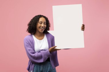 Neşeli genç bir Afrikalı Amerikalı kadın elinde boş bir konuşma balonu tutarken gülümsüyor. Pembe stüdyo geçmişi olan sıradan bir kıyafet içinde reklam tasarımı fikirleri için yer sunuyor..