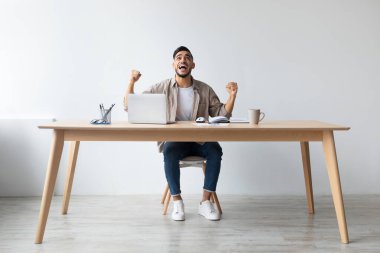 Bir Arap adam masasında oturuyor, heyecan verici haberleri okumak için dizüstü bilgisayar kullanıyor. O, başarısını yumruklarını sallayarak ve sevinçle bağırarak kutlar. Dekor minimalist bir ofis..