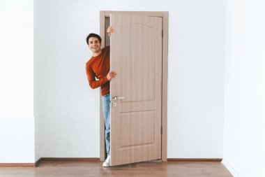 Bir adam parlak bir odada duruyor, açık ahşap bir kapıdan bakarken gülümsüyor. Duvarlar boş ve zemin ahşap. Ev dekorasyonu için mükemmel bir karşılama atmosferi yaratıyor..