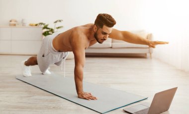 Odaklanmış bir adam, bir kolu uzatılmış bir şekilde bir dizüstü bilgisayarda çalışırken şınav çeker. Parlak odada modern bir tasarım ve arka planda yeşil bitkiler yer alıyor..